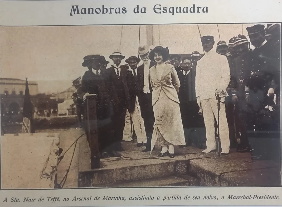 Recorte de imprensa intitulado “Manobras da Esquadra”: Nair de Teffé, de vestido claro e cabelo preso, em pé no cais do Arsenal de Marinha, cercada por homens de terno e chapéu e por oficiais de uniforme branco e escuro, com o casario do arsenal ao fundo
